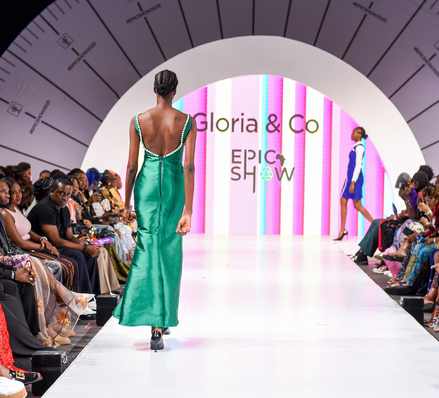 Model walking down a runway at a fashion show with 'Gloria & Co' branding.