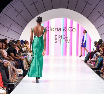 Model walking down a runway at a fashion show with 'Gloria & Co' branding.